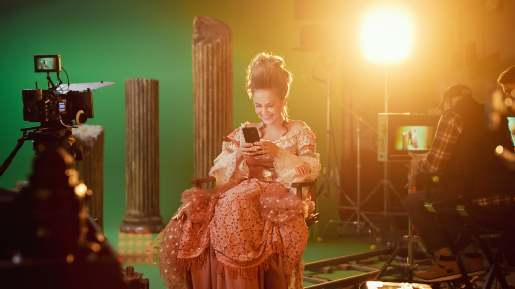 On Period Costume Drama Film Set: Beautiful Smiling Actress Wearing Renaissance Dress, Sitting on a Chair Using Smartphone with Green Screen in the Background. Studio Shooting Historical Blockbuster